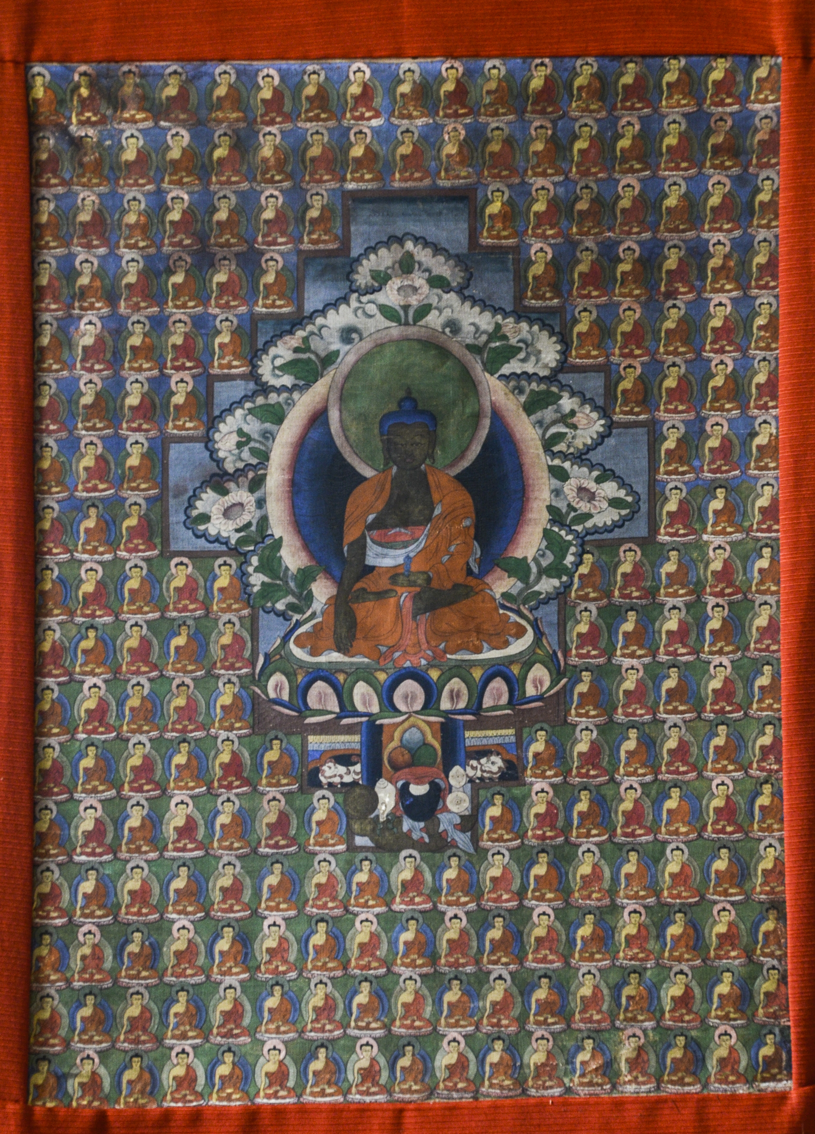 Pintura rectangular sobre tela típica de la zona de Tíbet. En el centro la figura del Buda Akṣobhya rodeado por muchas imágenes de budas más pequeños en tamaño.